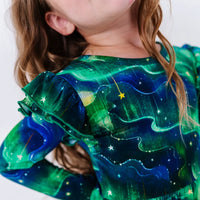 Close up of a flutter ruffle on the shoulder of a long sleeve twirl dress, showing a green and blue northern lights print with tiny stars.
