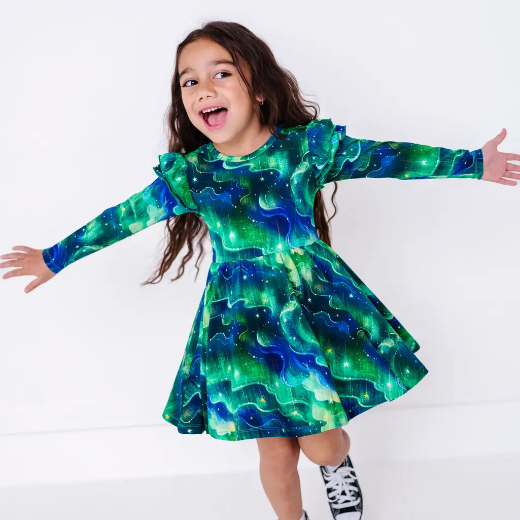A child twirling with arms out and a big smile as the skirt flares, in a long sleeve twirl dress with flutter ruffles at the shoulders in a green and blue northern lights print with tiny stars.