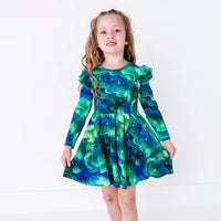 A child standing and holding out the skirt of a long sleeve twirl dress with flutter ruffles at the shoulders, in a green and blue northern lights print with tiny stars.