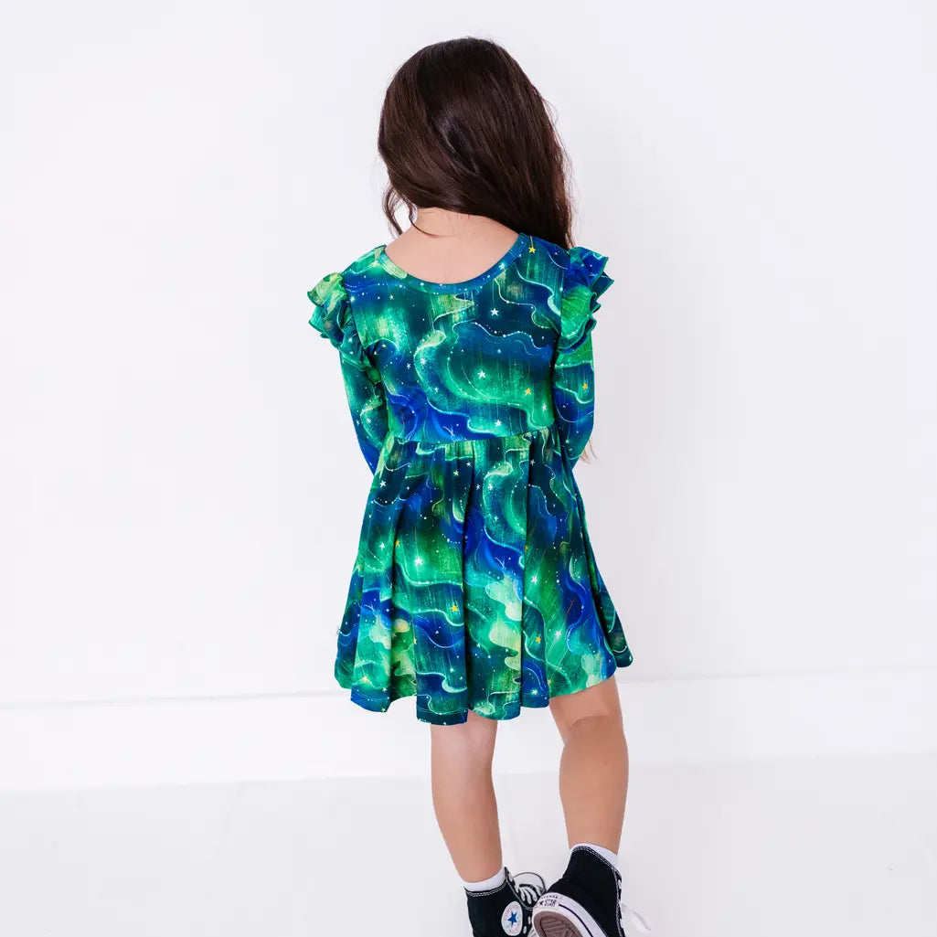 Back view of a child with long hair walking in a long sleeve twirl dress with flutter ruffles at the shoulders in a green and blue northern lights print with tiny stars.