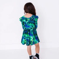 Back view of a child with long hair walking in a long sleeve twirl dress with flutter ruffles at the shoulders in a green and blue northern lights print with tiny stars.