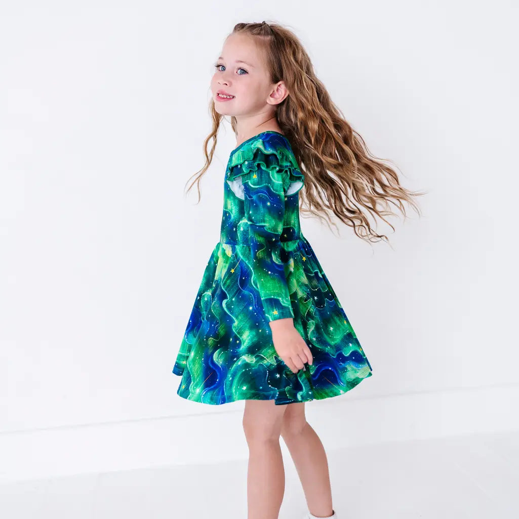 Side view of a smiling child twirling with long wavy hair flying, in a long sleeve twirl dress with flutter ruffles at the shoulders in a green and blue northern lights print with tiny stars.