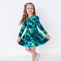 A smiling child stepping lightly and holding out the skirt of a long sleeve twirl dress with flutter ruffles at the shoulders, in a green and blue northern lights print with tiny stars.