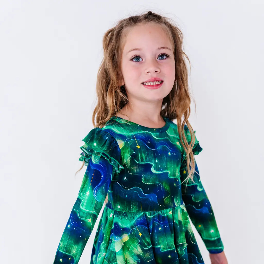 Close up portrait of a smiling child with wavy hair, in a long sleeve twirl dress with flutter ruffles at the shoulders in a green and blue northern lights print with tiny stars.