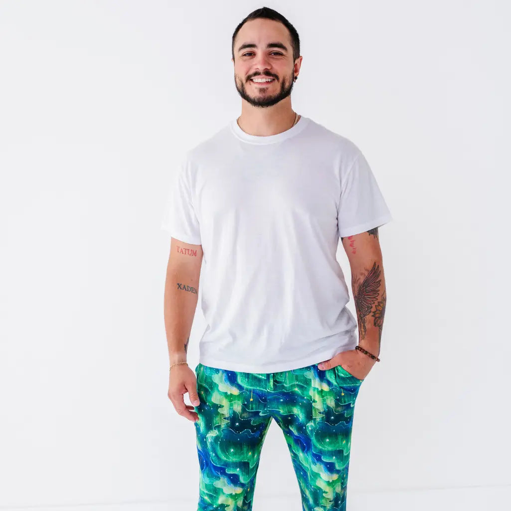 A smiling man in a white tee wearing pajama pants with a green and blue northern lights pattern.
