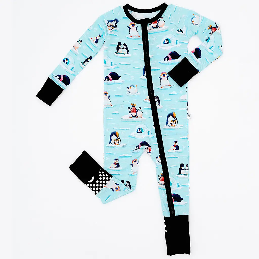 Flat lay of a zip up convertible baby pajama with black trim and cuffs in a light blue print of penguins playing on ice floes on a white background.