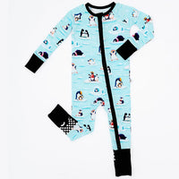 Flat lay of a zip up convertible baby pajama with black trim and cuffs in a light blue print of penguins playing on ice floes on a white background.