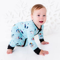 A smiling baby crawling on a white floor and looking at the camera, wearing a zip up pajama with black trim and cuffs in a light blue print of penguins playing on ice floes.