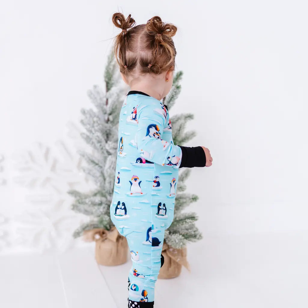 Back view of a toddler standing next to a small snowy tree, wearing a zip up pajama with black trim and cuffs in a light blue print of penguins playing on ice floes.