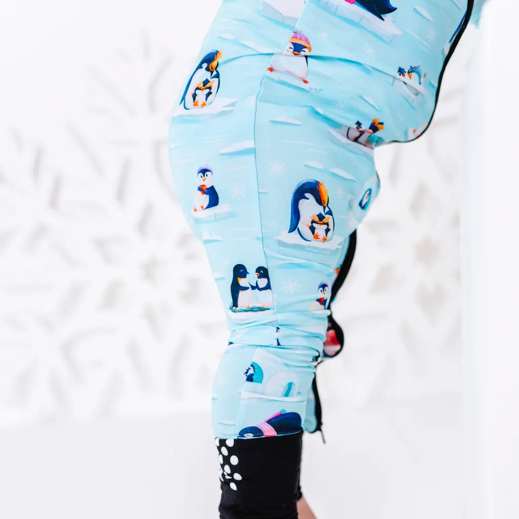 Close up of the side of the leg and the foot cuff with grip dots of a zip up baby pajama with black trim in a light blue print of penguins playing on ice floes.