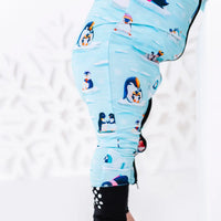 Close up of the side of the leg and the foot cuff with grip dots of a zip up baby pajama with black trim in a light blue print of penguins playing on ice floes.