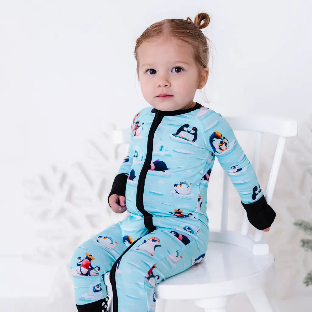 A toddler sitting on a small white chair and looking at the camera, wearing a zip up pajama with black trim and cuffs in a light blue print of penguins playing on ice floes.