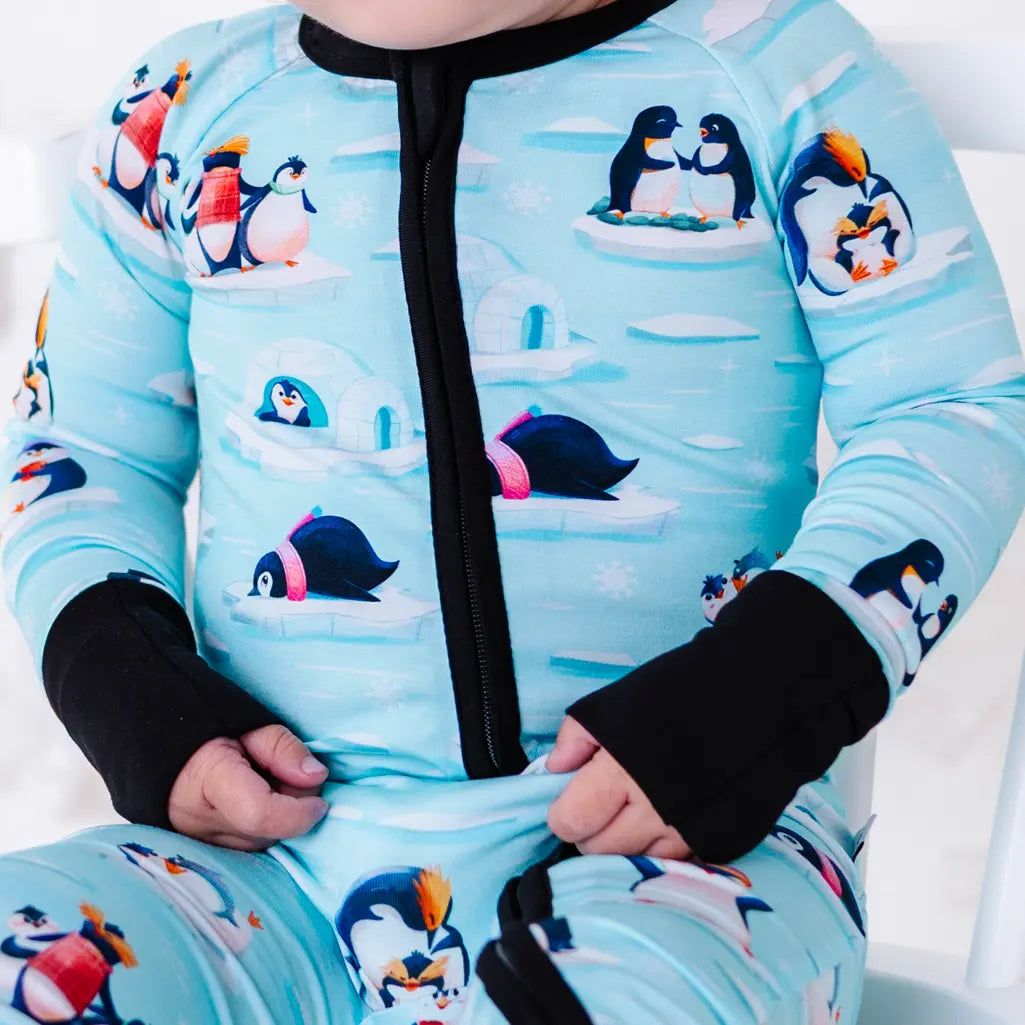 Close up of the front zipper and chest of a zip up baby pajama with black trim and cuffs in a light blue print of penguins playing on ice floes.