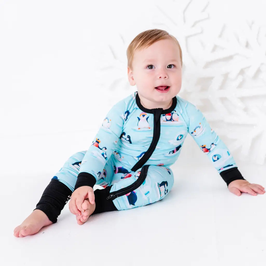 A baby sitting on a white floor with one bare foot showing and looking at the camera, wearing a zip up pajama with black trim and cuffs in a light blue print of penguins playing on ice floes.