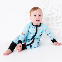 A baby sitting on a white floor with one bare foot showing and looking at the camera, wearing a zip up pajama with black trim and cuffs in a light blue print of penguins playing on ice floes.