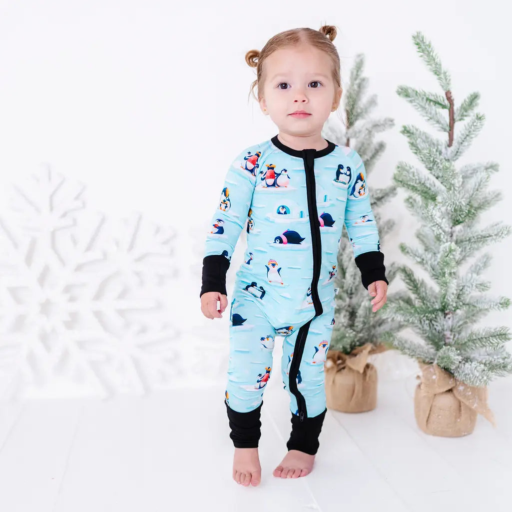 A toddler standing barefoot next to two small snowy trees and looking at the camera, wearing a zip up pajama with black trim and cuffs in a light blue print of penguins playing on ice floes.