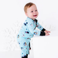 A smiling baby standing and leaning on a white block, wearing a zip up pajama with black trim and cuffs in a light blue print of penguins playing on ice floes.