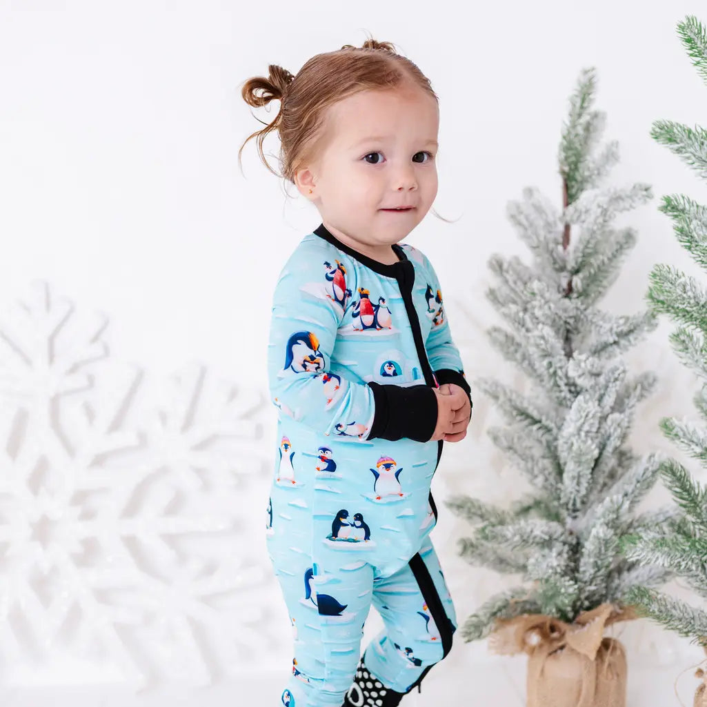 A toddler standing next to a small snowy tree and looking at the camera, wearing a zip up pajama with black trim and cuffs in a light blue print of penguins playing on ice floes.
