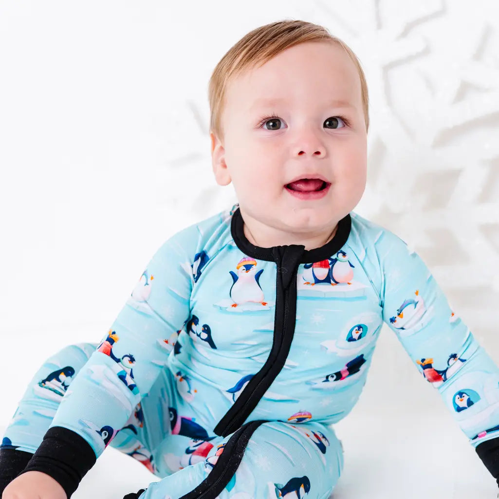 Close portrait of a baby with an open mouth smile, wearing a zip up pajama with black trim and cuffs in a light blue print of penguins playing on ice floes.