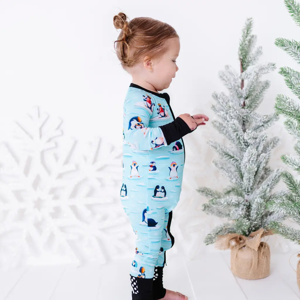 Side view of a toddler standing in profile with one hand out in front, wearing a zip up pajama with black trim and cuffs in a light blue print of penguins playing on ice floes.