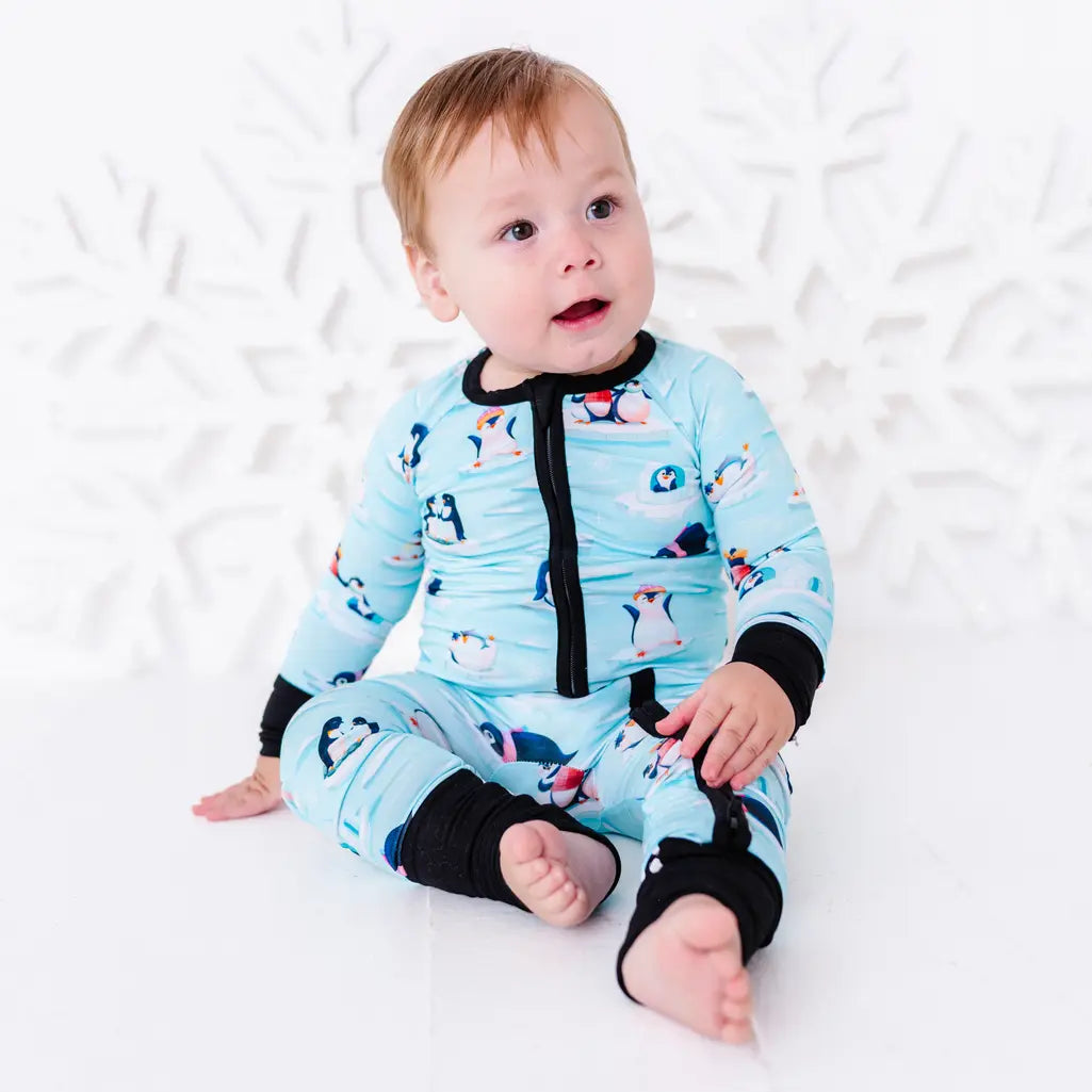 A baby sitting barefoot on a white floor and looking up with an open mouth, wearing a zip up pajama with black trim and cuffs in a light blue print of penguins playing on ice floes.