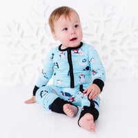 A baby sitting barefoot on a white floor and looking up with an open mouth, wearing a zip up pajama with black trim and cuffs in a light blue print of penguins playing on ice floes.