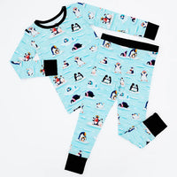 Flat lay of a long sleeve pajama top and matching pants with black cuffs in a light blue print of penguins playing on ice floes on a white background.