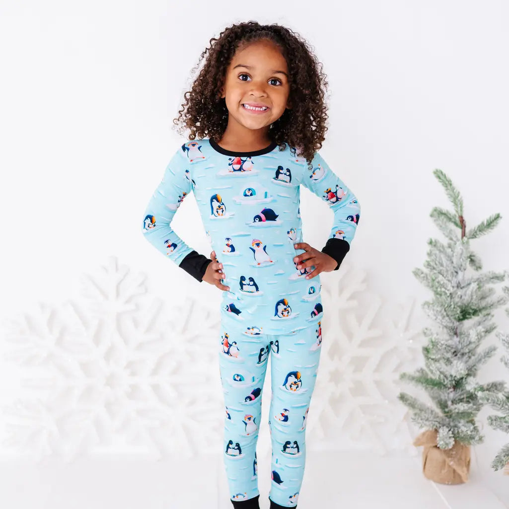 A smiling child standing with both hands on the hips next to a small snowy tree, wearing a long sleeve pajama set with black cuffs in a light blue print of penguins playing on ice floes.