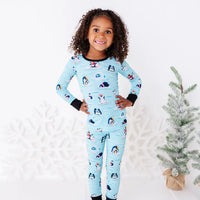 A smiling child standing with both hands on the hips next to a small snowy tree, wearing a long sleeve pajama set with black cuffs in a light blue print of penguins playing on ice floes.