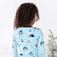 Back view of a child with curly hair, wearing a long sleeve pajama top with black cuffs in a light blue print of penguins playing on ice floes.