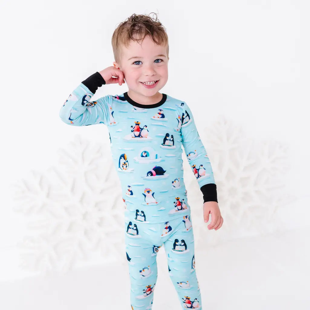 A smiling toddler with one hand raised to the head, wearing a long sleeve pajama set with black cuffs in a light blue print of penguins playing on ice floes.