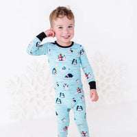 A smiling toddler with one hand raised to the head, wearing a long sleeve pajama set with black cuffs in a light blue print of penguins playing on ice floes.