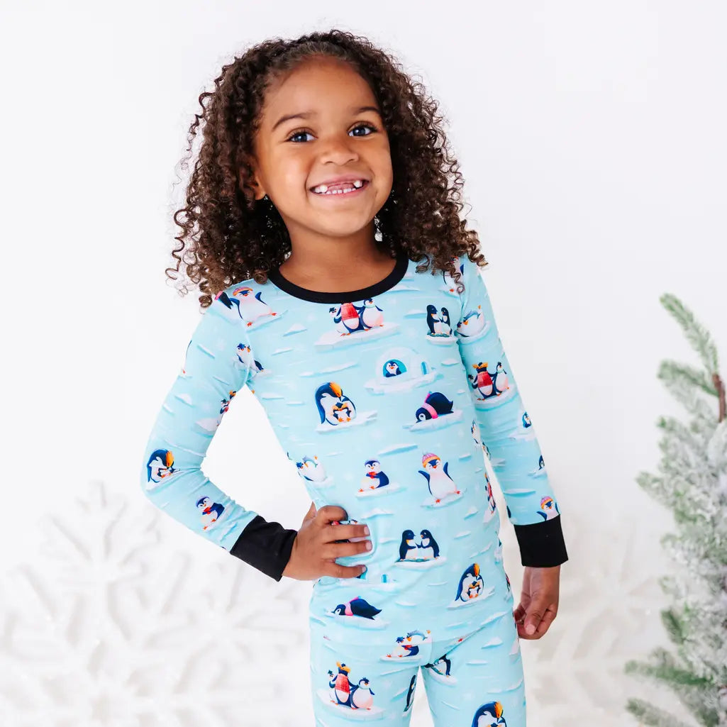A child with a big smile and one hand on the hip, shown from the waist up, wearing a long sleeve pajama top with black cuffs in a light blue print of penguins playing on ice floes.