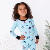 A child with a big smile and one hand on the hip, shown from the waist up, wearing a long sleeve pajama top with black cuffs in a light blue print of penguins playing on ice floes.