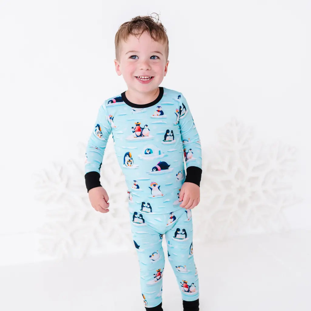A smiling toddler standing with arms at the sides, wearing a long sleeve pajama set with black cuffs in a light blue print of penguins playing on ice floes.