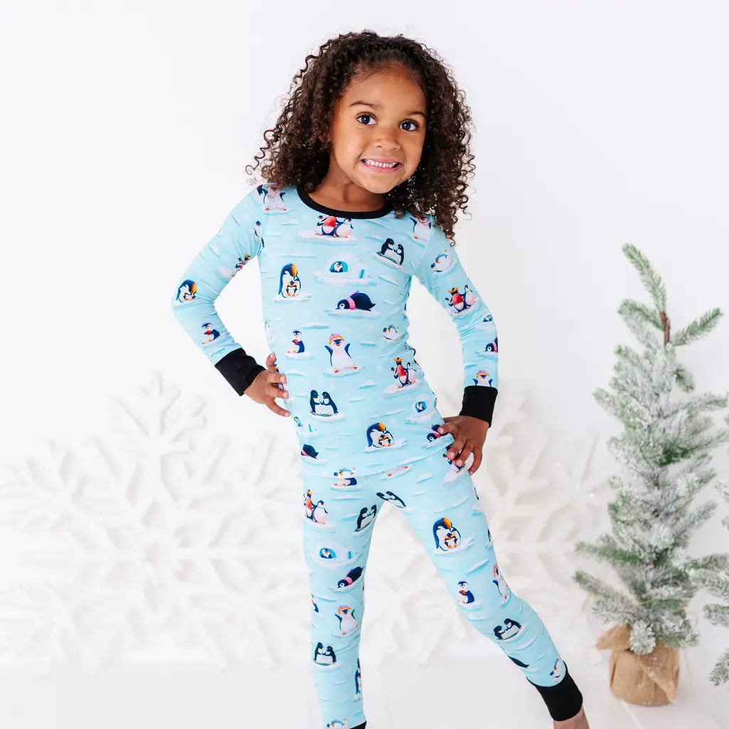A smiling child standing with one hand on the hip and one leg bent, wearing a long sleeve pajama set with black cuffs in a light blue print of penguins playing on ice floes.