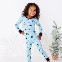 A smiling child standing with one hand on the hip and one leg bent, wearing a long sleeve pajama set with black cuffs in a light blue print of penguins playing on ice floes.