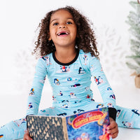 A child laughing and looking up while sitting on the floor with a board book, wearing a long sleeve pajama set with black cuffs in a light blue print of penguins playing on ice floes.