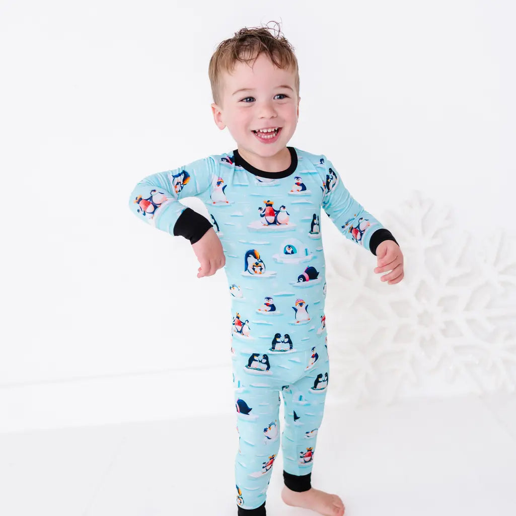 A smiling toddler standing barefoot with one arm swinging out, wearing a long sleeve pajama set with black cuffs in a light blue print of penguins playing on ice floes.