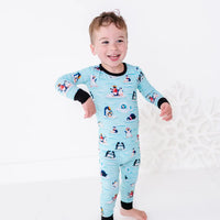 A smiling toddler standing barefoot with one arm swinging out, wearing a long sleeve pajama set with black cuffs in a light blue print of penguins playing on ice floes.