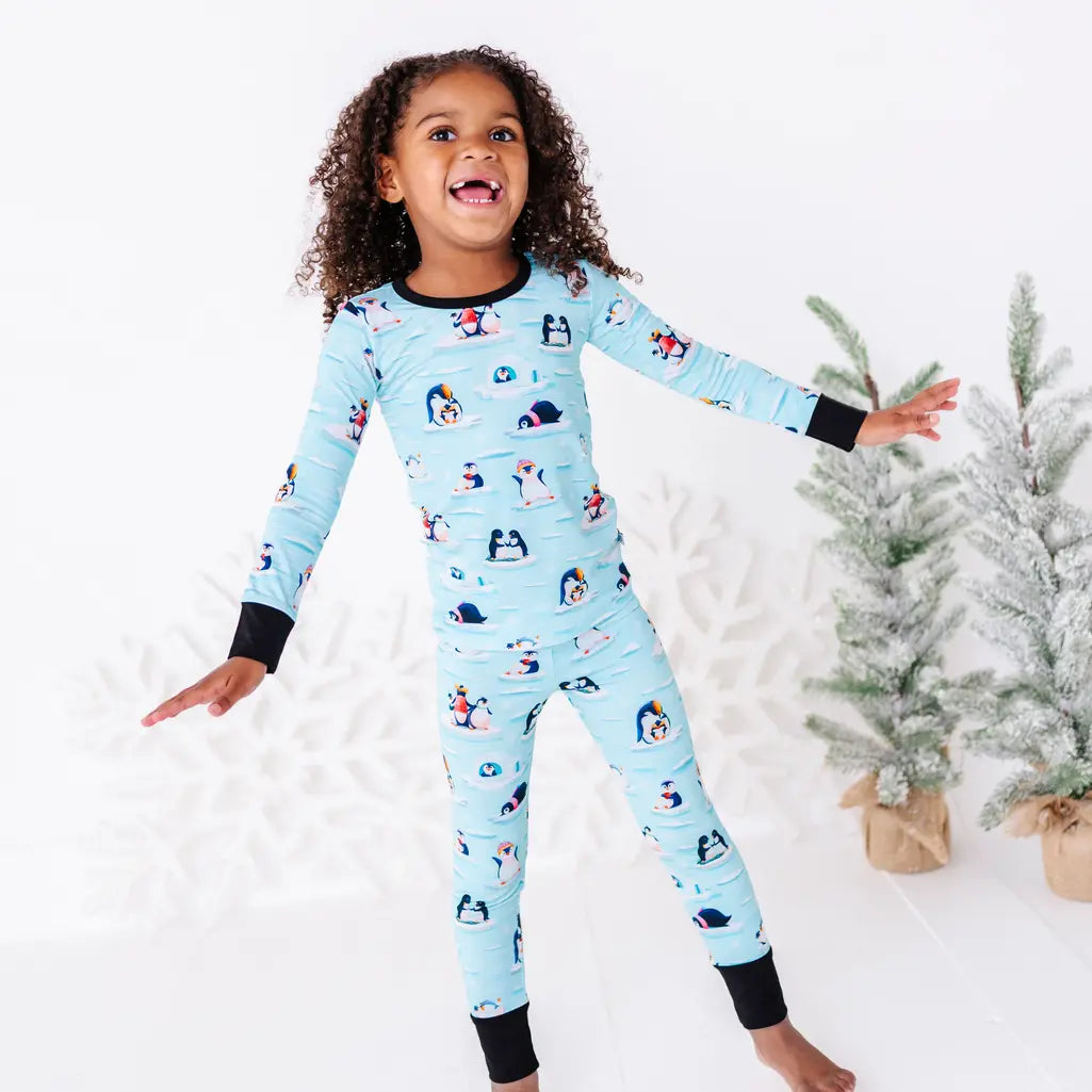 A child laughing with arms stretched out wide next to two small snowy trees, wearing a long sleeve pajama set with black cuffs in a light blue print of penguins playing on ice floes.