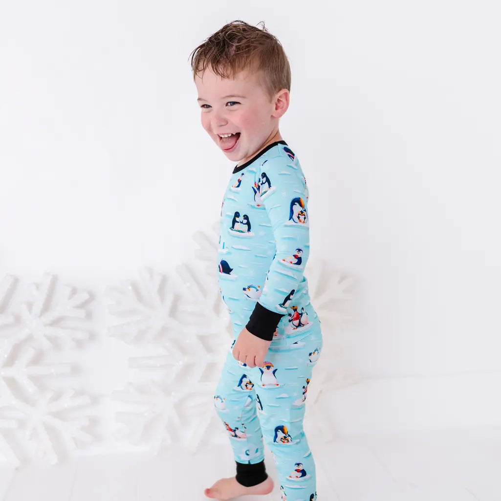 Side view of a laughing toddler standing barefoot, wearing a long sleeve pajama set with black cuffs in a light blue print of penguins playing on ice floes.