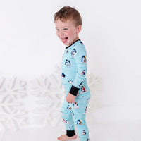 Side view of a laughing toddler standing barefoot, wearing a long sleeve pajama set with black cuffs in a light blue print of penguins playing on ice floes.