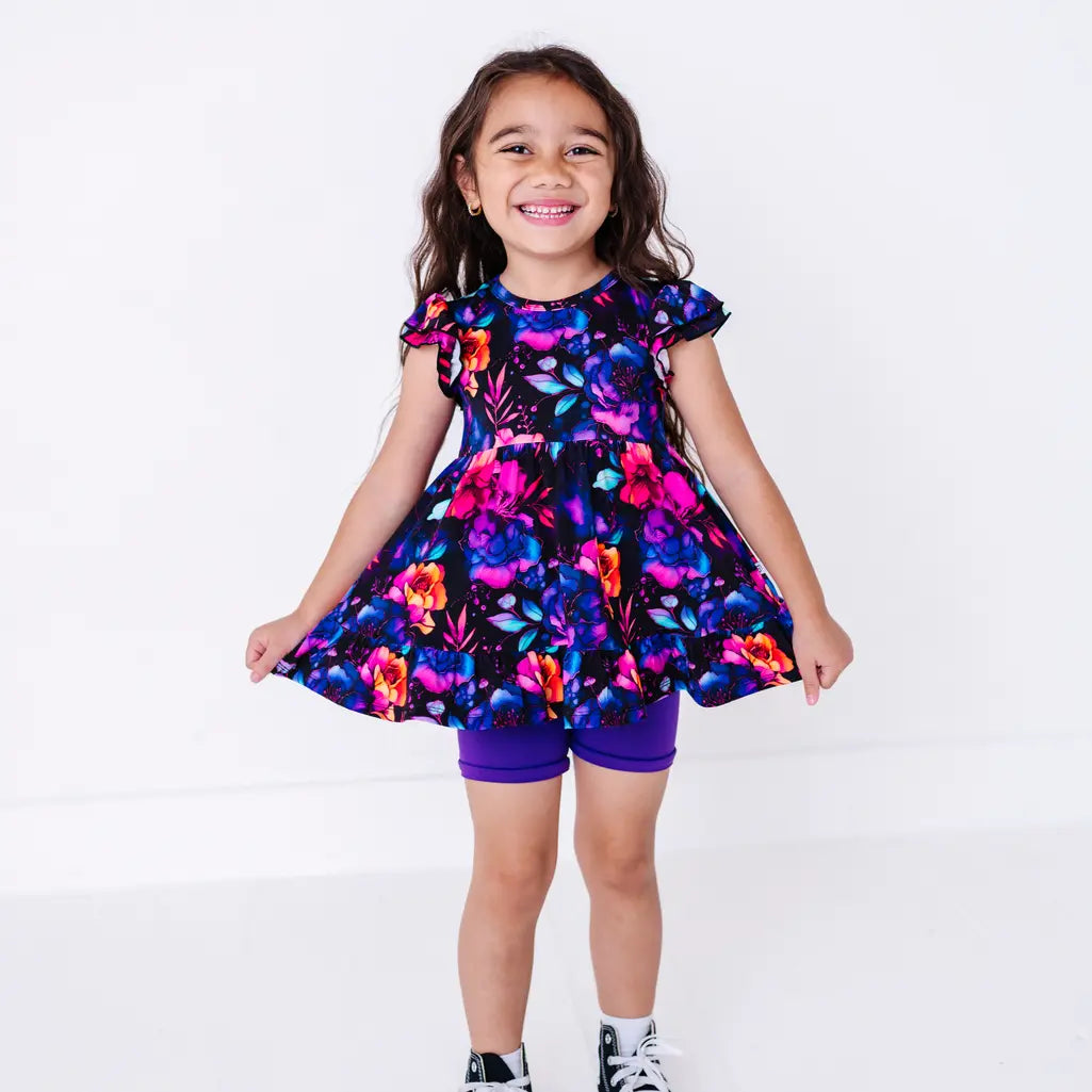 A child with long hair grinning and holding out the hem of a flutter sleeve peplum top in a black watercolor floral print with purple bike shorts.