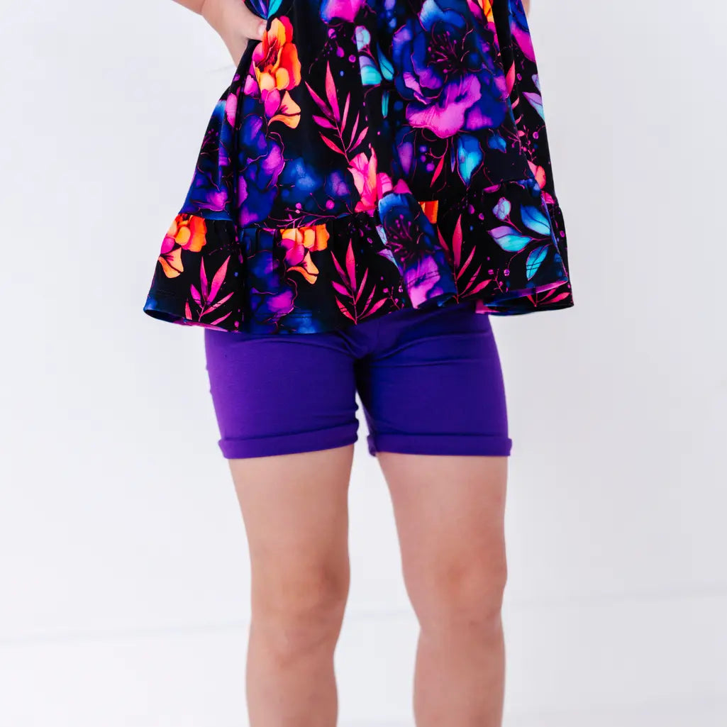 Close up of the ruffled hem of a flutter sleeve peplum top in a black watercolor floral print over purple bike shorts.