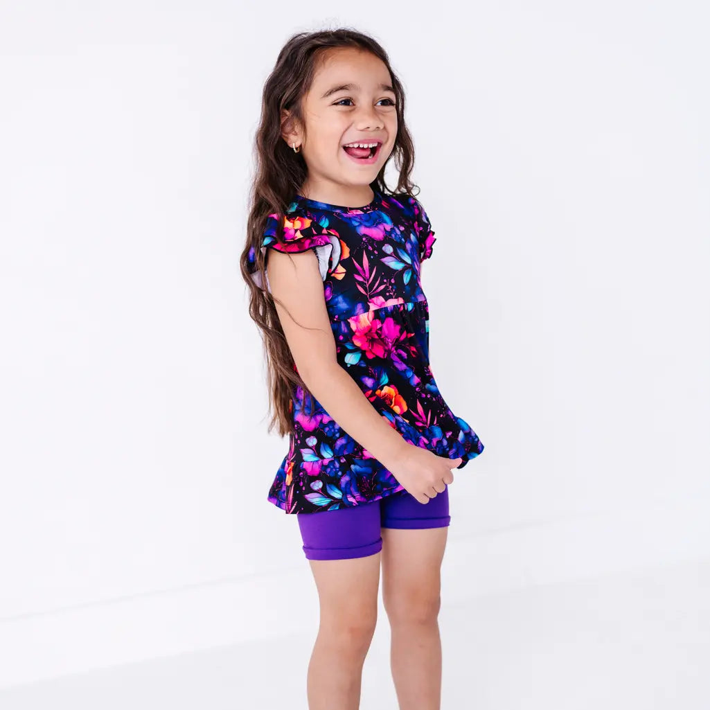 Side view of a laughing child looking off to the side, in a flutter sleeve peplum top in a black watercolor floral print with purple bike shorts.