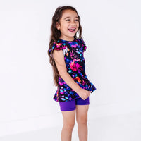 Side view of a laughing child looking off to the side, in a flutter sleeve peplum top in a black watercolor floral print with purple bike shorts.