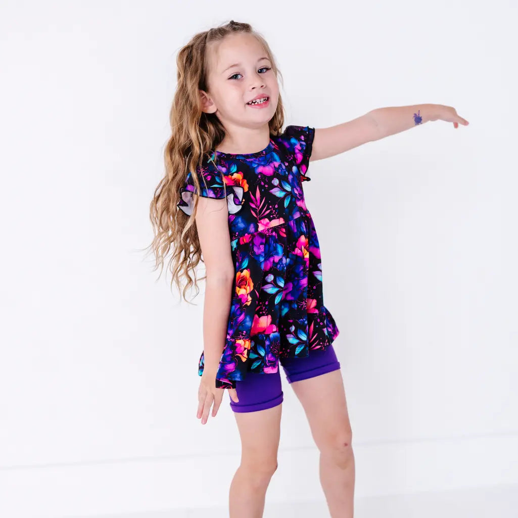 A child smiling with one arm stretched out to the side, in a flutter sleeve peplum top in a black watercolor floral print with purple bike shorts.