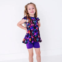 A child smiling with both hands on the hips, in a flutter sleeve peplum top in a black watercolor floral print with purple bike shorts.