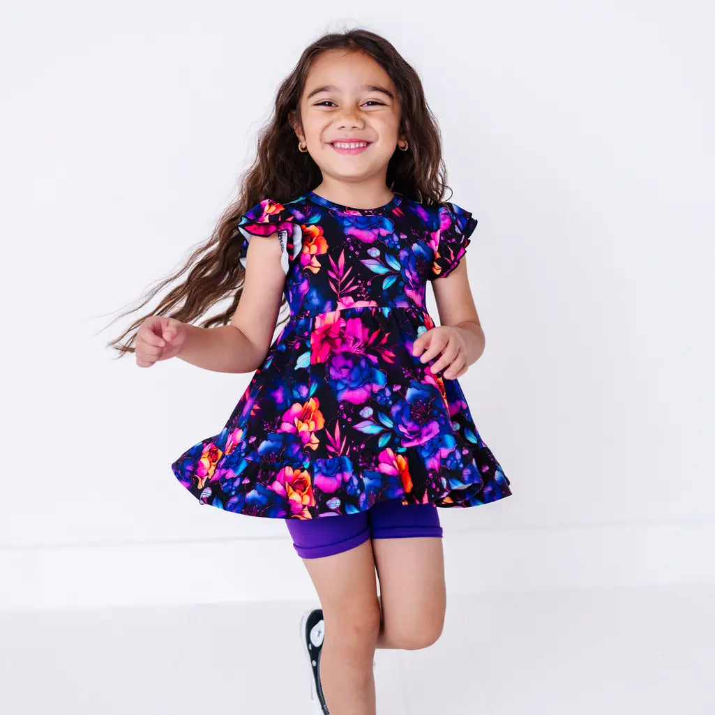 A smiling child jumping with long hair flying, in a flutter sleeve peplum top in a black watercolor floral print with purple bike shorts.